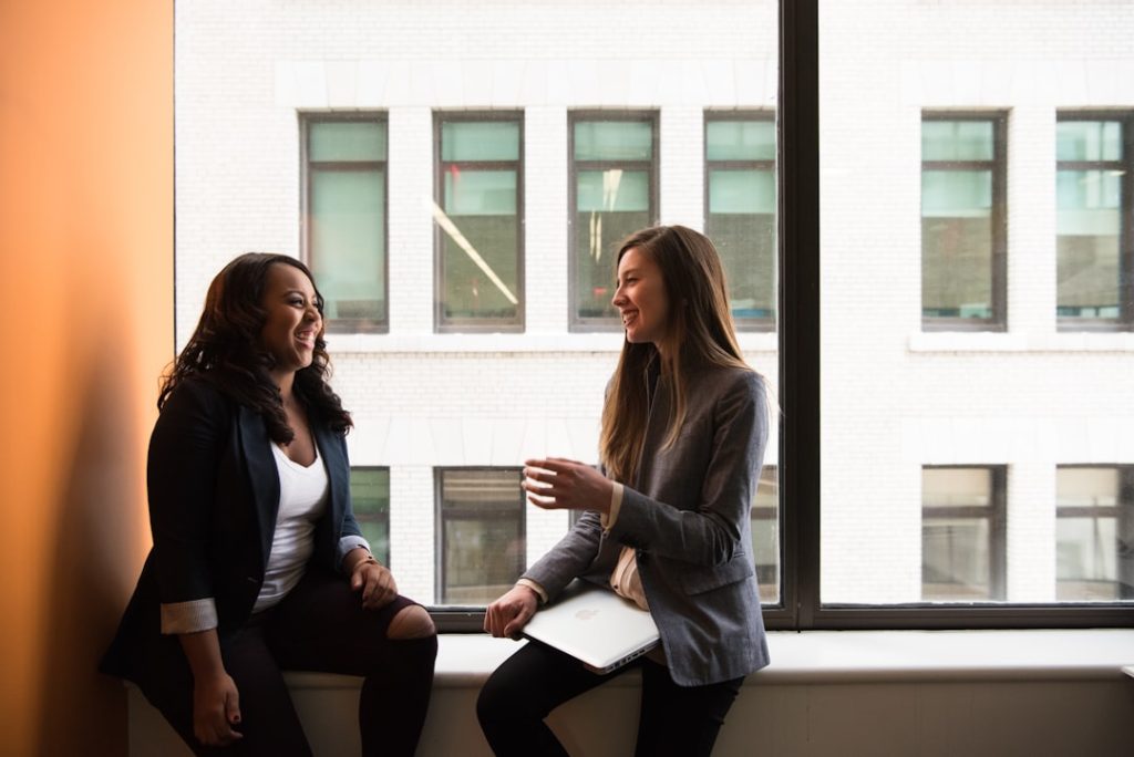 Two women sitting by a window in conversation, one coaching the other, the kind of one-on-one session coaching software is meant to support