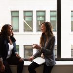 Two women sitting by a window in conversation, one coaching the other, the kind of one-on-one session coaching software is meant to support