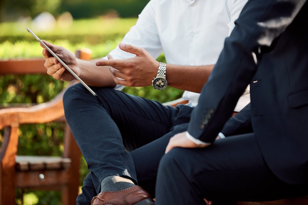 Two people sitting side by side outdoors in a one-on-one conversation, one holding a tablet and gesturing while explaining