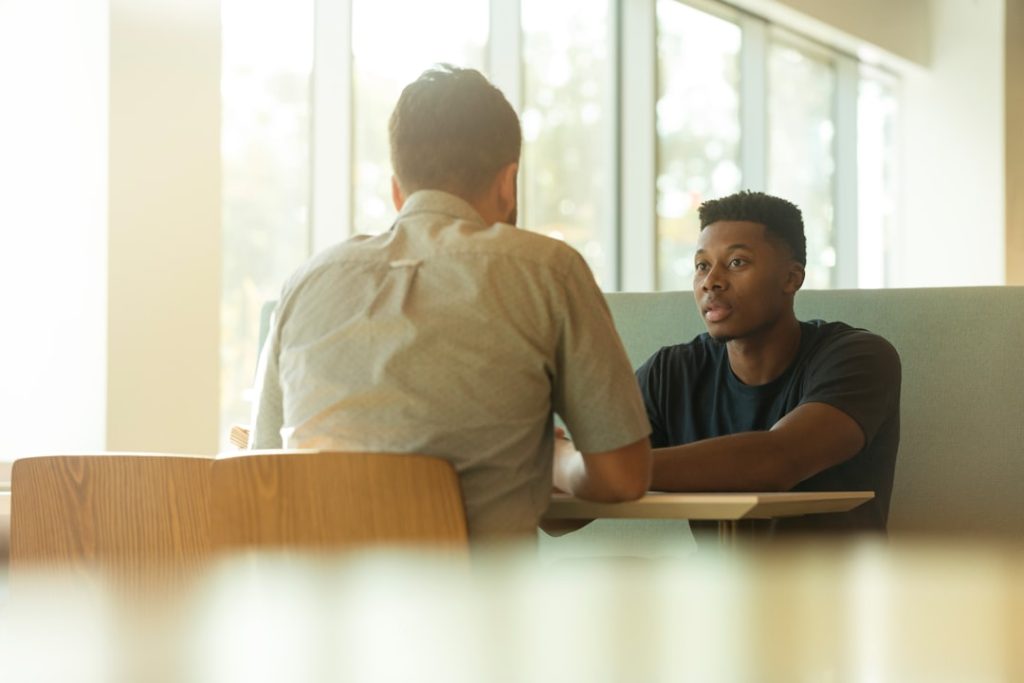 Two people leaning in mid-conversation across a sunlit cafe table, the kind of one-on-one client conversation that reveals a coaching niche.