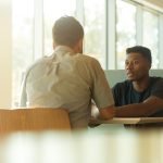 Two people leaning in mid-conversation across a sunlit cafe table, the kind of one-on-one client conversation that reveals a coaching niche.