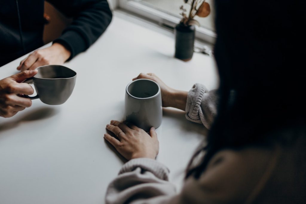 Two people sitting across a small table holding coffee mugs mid-conversation, the kind of one-on-one talk that brings a new coaching business its first clients.