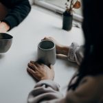 Two people sitting across a small table holding coffee mugs mid-conversation, the kind of one-on-one talk that brings a new coaching business its first clients.