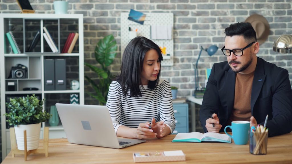 A coach and a client sitting side by side at a table, reviewing progress on a laptop during a one-on-one accountability check-in.