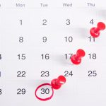 A white monthly calendar page shot from above, with red push pins stuck through four dates in a diagonal line and the 30th circled in red marker.