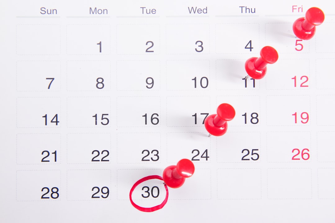 A white monthly calendar page shot from above, with red push pins stuck through four dates in a diagonal line and the 30th circled in red marker.