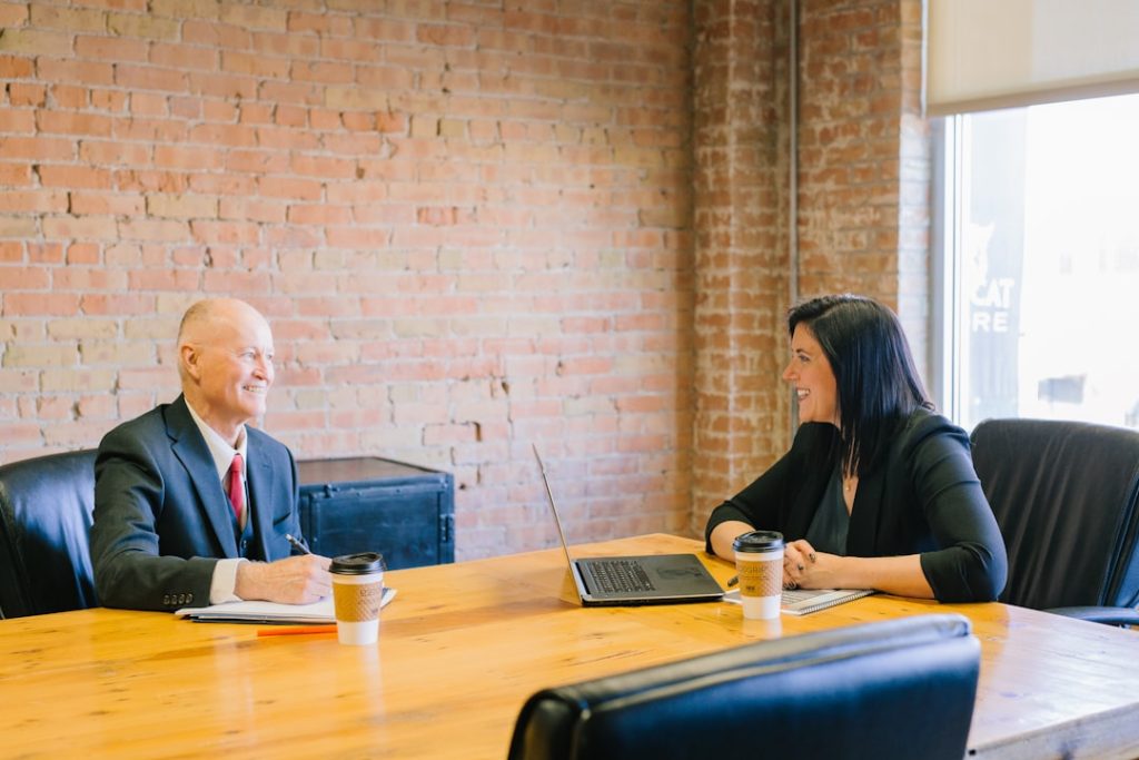 A coach and a client sit across a wooden table in a brick-walled office, talking and smiling, with an open notebook, a pen, a laptop and two coffee cups between them.