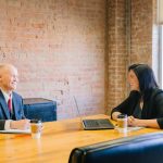 A coach and a client sit across a wooden table in a brick-walled office, talking and smiling, with an open notebook, a pen, a laptop and two coffee cups between them.