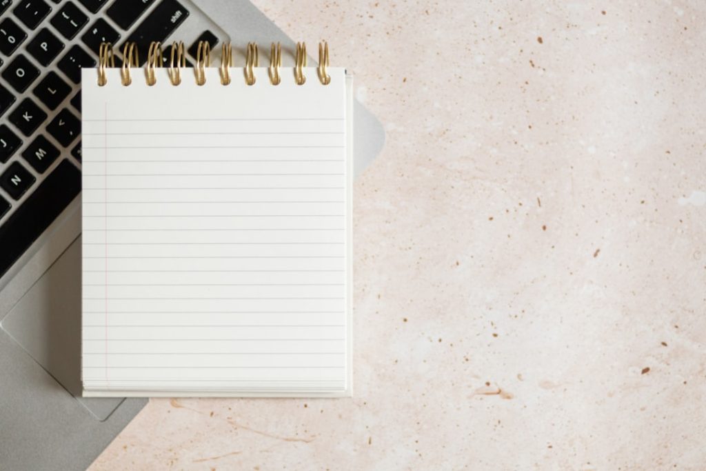 A blank spiral-bound lined notepad resting on an open laptop keyboard on a pale speckled stone desk, with open space beside it.