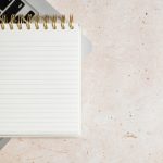 A blank spiral-bound lined notepad resting on an open laptop keyboard on a pale speckled stone desk, with open space beside it.