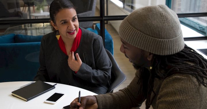 Two people sitting across a round white table in a bright office, mid-conversation, with notebooks, a phone and a pen in front of them.