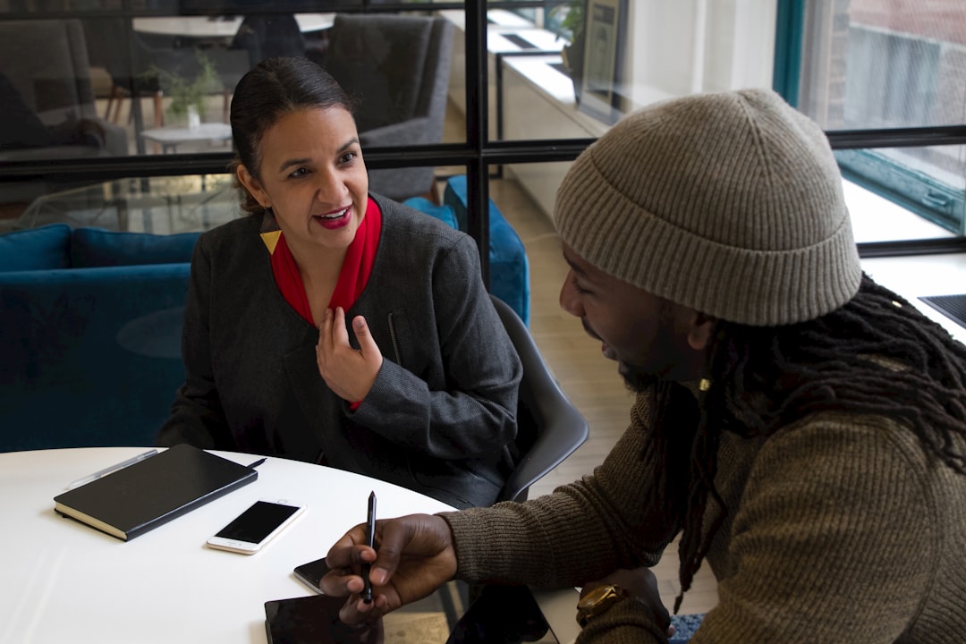Two people sitting across a round white table in a bright office, mid-conversation, with notebooks, a phone and a pen in front of them.