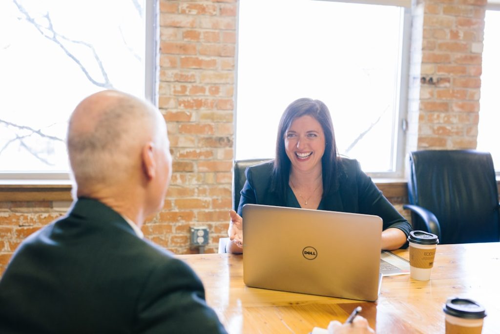 A smiling woman in a dark blazer talks with a man seated across a wooden table in a brick-walled office, an open laptop and coffee cups between them.