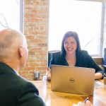 A smiling woman in a dark blazer talks with a man seated across a wooden table in a brick-walled office, an open laptop and coffee cups between them.