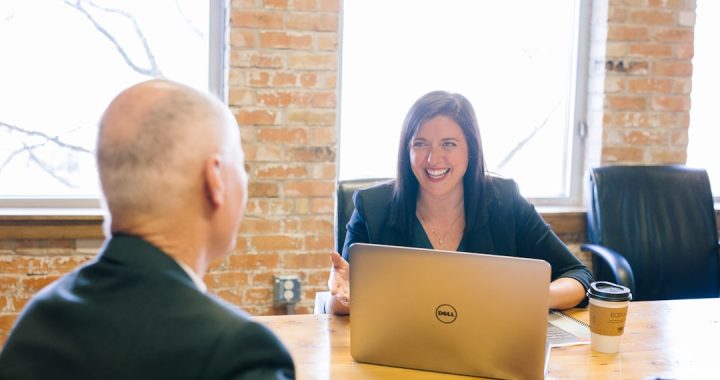 A smiling woman in a dark blazer talks with a man seated across a wooden table in a brick-walled office, an open laptop and coffee cups between them.