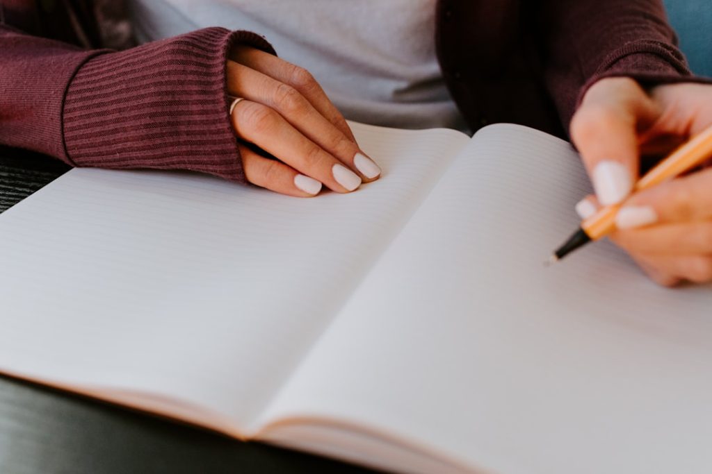 Close-up of a person in a maroon sweater resting one hand on an open blank ruled notebook while holding a pen ready to write on the facing page.