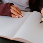 Close-up of a person in a maroon sweater resting one hand on an open blank ruled notebook while holding a pen ready to write on the facing page.
