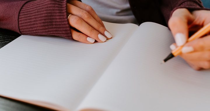 Close-up of a person in a maroon sweater resting one hand on an open blank ruled notebook while holding a pen ready to write on the facing page.