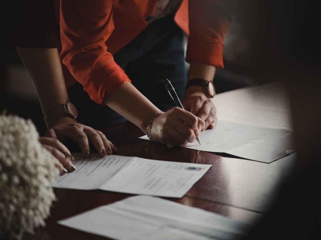 Two people lean over a polished wooden table filling out printed forms, the one in an orange blazer signing a page with a pen while several other completed documents lie spread across the surface.