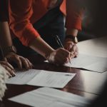 Two people lean over a polished wooden table filling out printed forms, the one in an orange blazer signing a page with a pen while several other completed documents lie spread across the surface.