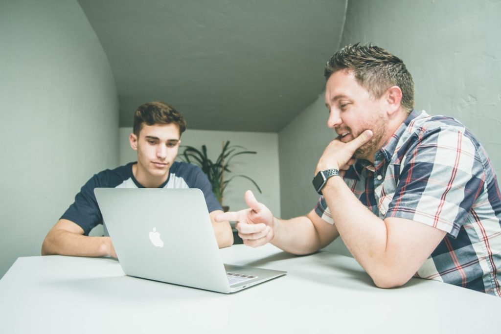 Two people sit across a white table in a bright room, one gesturing toward an open laptop while the other looks at the screen, in a one-on-one coaching conversation.