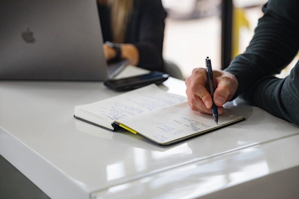 A coach and client sitting across a table, one of them writing a coaching plan by hand in an open notebook beside an open laptop.