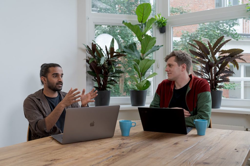 Two people sitting side by side at a wooden table in a bright, plant-filled office, one gesturing mid-sentence while the other listens, with open laptops and coffee mugs between them.