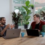 Two people sitting side by side at a wooden table in a bright, plant-filled office, one gesturing mid-sentence while the other listens, with open laptops and coffee mugs between them.