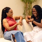 Two people sitting on a white couch in a warmly lit room, turned toward each other mid-conversation and laughing, with a potted plant behind them.
