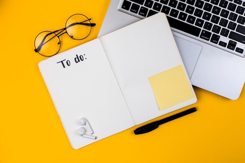 Flat lay on a yellow background: an open notebook with blank dotted pages and the handwritten heading "To do:", a black pen, a blank yellow sticky note, round eyeglasses and wireless earbuds beside a laptop keyboard.