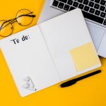 Flat lay on a yellow background: an open notebook with blank dotted pages and the handwritten heading "To do:", a black pen, a blank yellow sticky note, round eyeglasses and wireless earbuds beside a laptop keyboard.