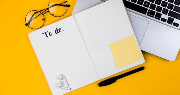 Flat lay on a yellow background: an open notebook with blank dotted pages and the handwritten heading "To do:", a black pen, a blank yellow sticky note, round eyeglasses and wireless earbuds beside a laptop keyboard.