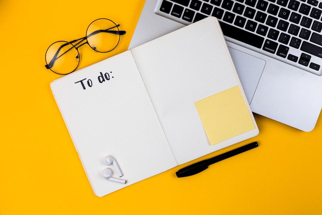 Flat lay on a yellow background: an open notebook with blank dotted pages and the handwritten heading "To do:", a black pen, a blank yellow sticky note, round eyeglasses and wireless earbuds beside a laptop keyboard.