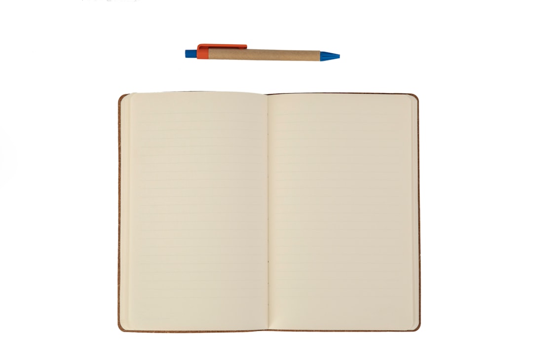 An open blank notebook with cream lined pages lying flat, a kraft-paper ballpoint pen with blue and orange trim resting above it, on a plain white background.