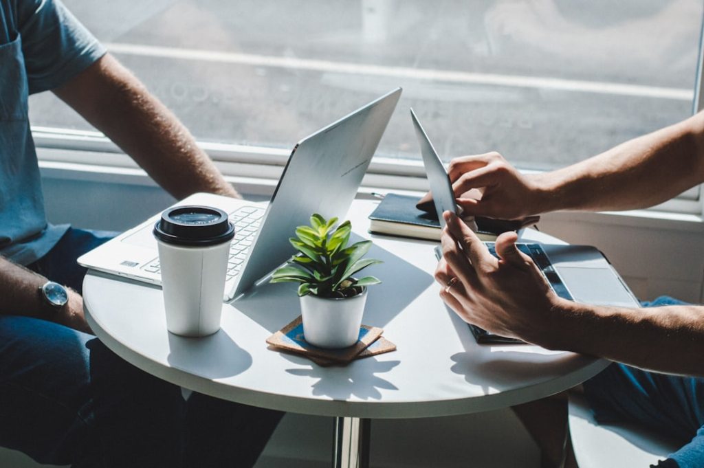 Two people sitting across a small round table beside a window, each with an open laptop, a takeaway coffee cup and a small potted plant between them, working through something together in a one-on-one meeting.