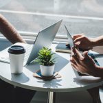 Two people sitting across a small round table beside a window, each with an open laptop, a takeaway coffee cup and a small potted plant between them, working through something together in a one-on-one meeting.