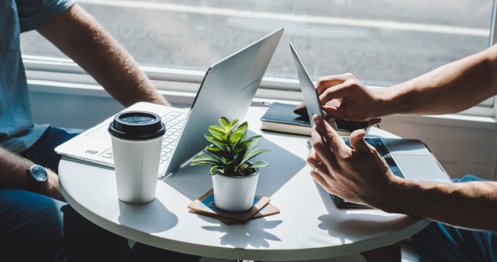 Two people sitting across a small round table beside a window, each with an open laptop, a takeaway coffee cup and a small potted plant between them, working through something together in a one-on-one meeting.