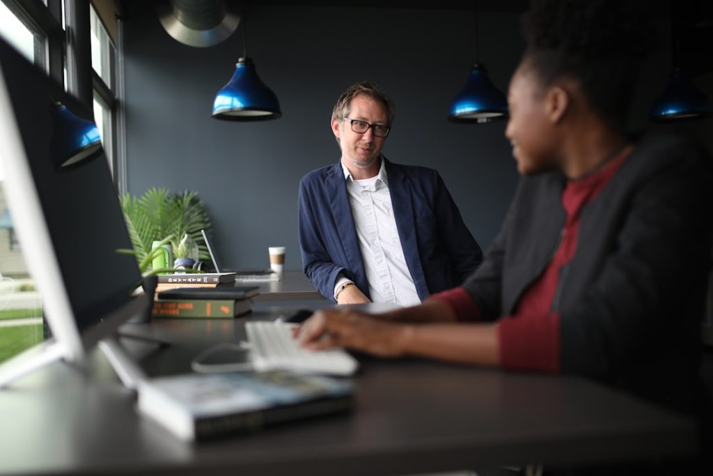Two colleagues talking across a desk in a dark-walled office with blue pendant lights, one seated at a computer with a keyboard and a stack of books, the other standing and listening.