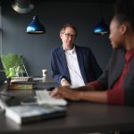 Two colleagues talking across a desk in a dark-walled office with blue pendant lights, one seated at a computer with a keyboard and a stack of books, the other standing and listening.