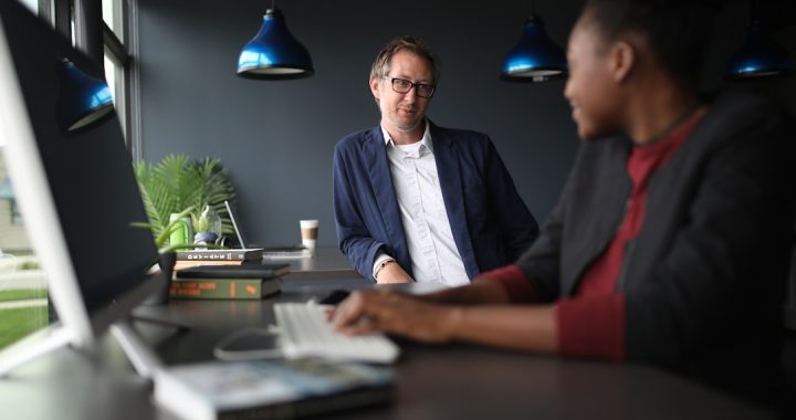 Two colleagues talking across a desk in a dark-walled office with blue pendant lights, one seated at a computer with a keyboard and a stack of books, the other standing and listening.
