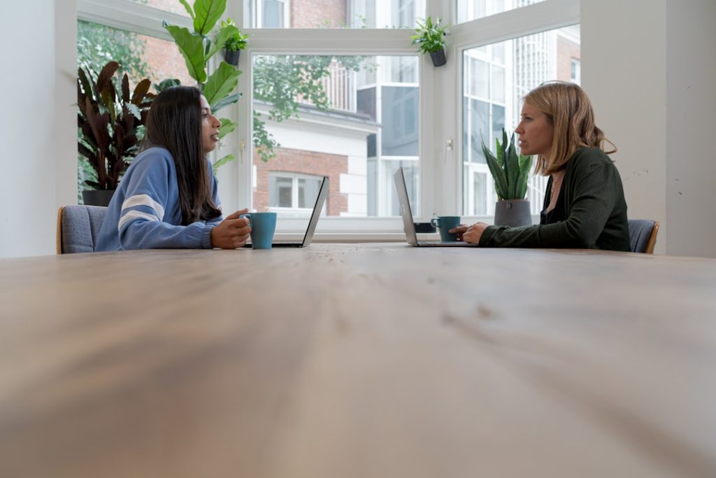 Two people sit across a long wooden table in a bright, plant-filled room, turned toward each other mid-conversation, with open laptops and mugs of coffee in front of them.