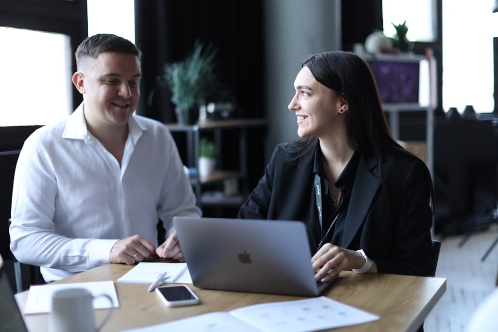 Two professionals sitting side by side at a wooden table in an office, turned toward each other mid-conversation, with an open laptop, a notebook and printed pages on the desk between them.
