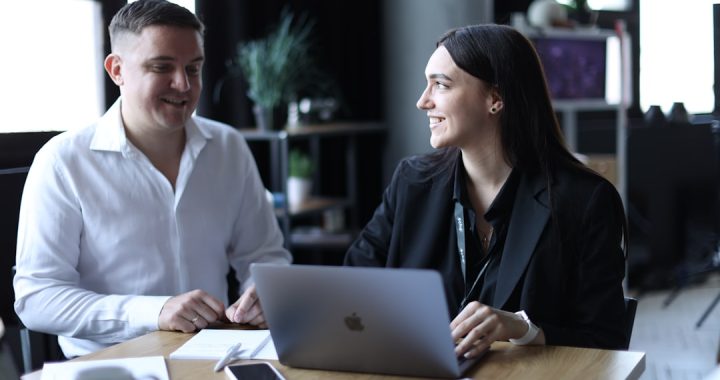Two professionals sitting side by side at a wooden table in an office, turned toward each other mid-conversation, with an open laptop, a notebook and printed pages on the desk between them.