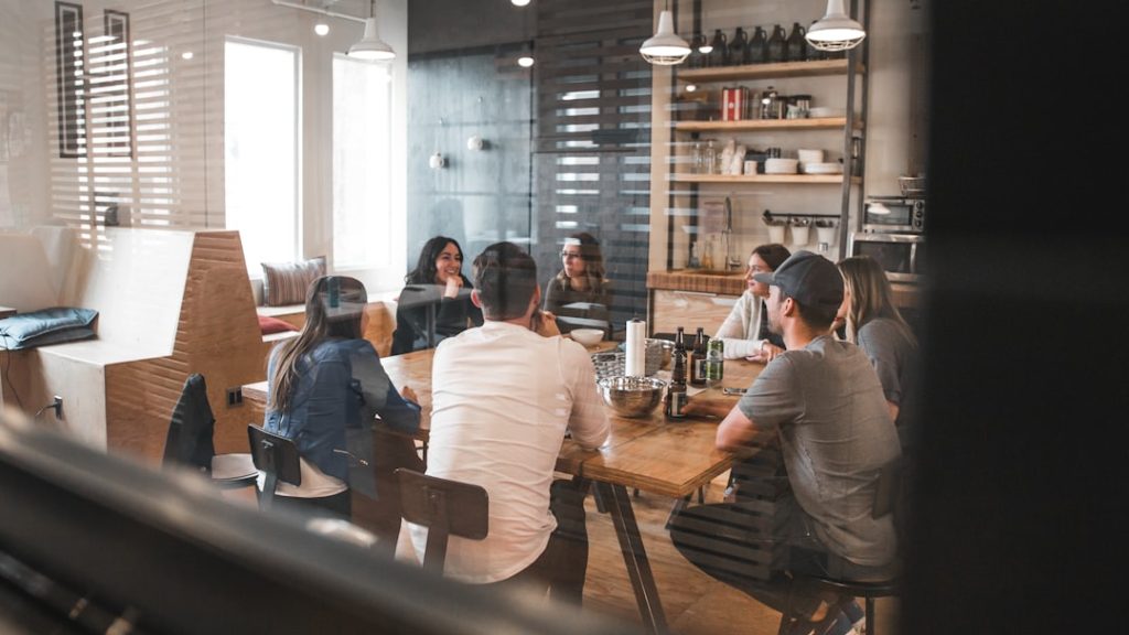 A group of about eight people seated around a long wooden table in a bright cafe workspace, mid-conversation during a group session