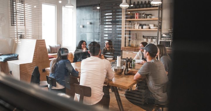 A group of about eight people seated around a long wooden table in a bright cafe workspace, mid-conversation during a group session