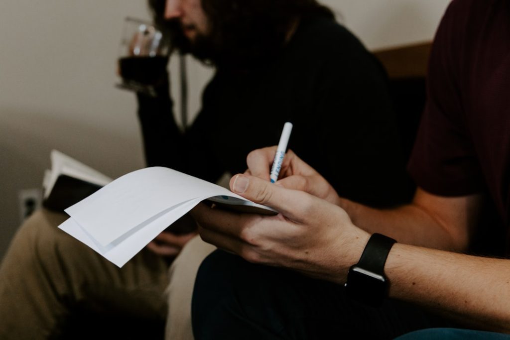 Two people seated side by side in a one-on-one conversation, one writing notes on a sheet of paper with a pen while the other listens holding a drink.