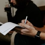 Two people seated side by side in a one-on-one conversation, one writing notes on a sheet of paper with a pen while the other listens holding a drink.