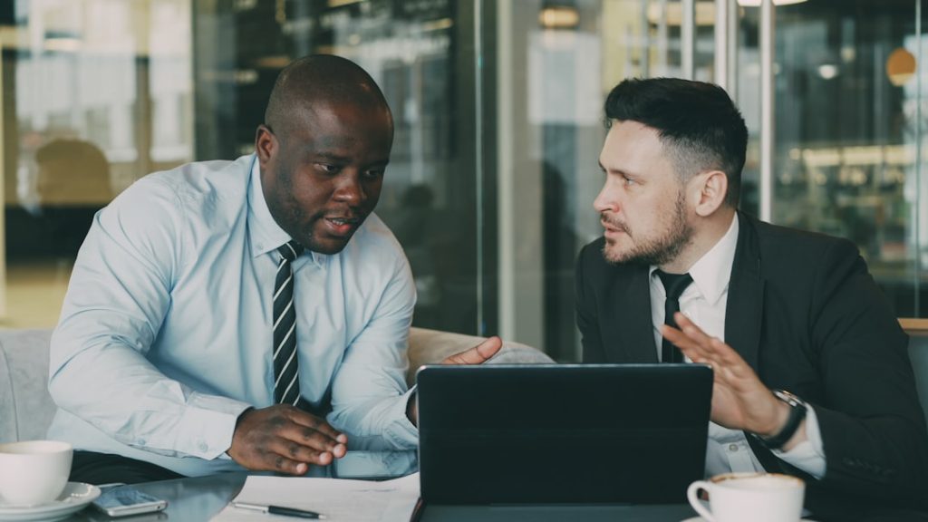 Two people in business attire sit side by side at a table in a glass-walled office, mid-conversation over an open laptop, with coffee cups and a notepad in front of them. One gestures while making a point and the other listens.