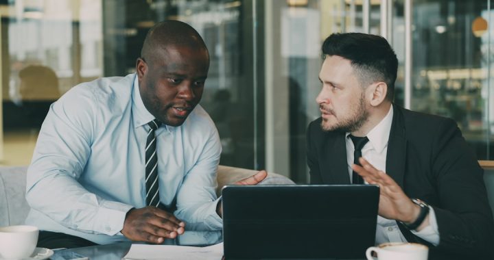 Two people in business attire sit side by side at a table in a glass-walled office, mid-conversation over an open laptop, with coffee cups and a notepad in front of them. One gestures while making a point and the other listens.