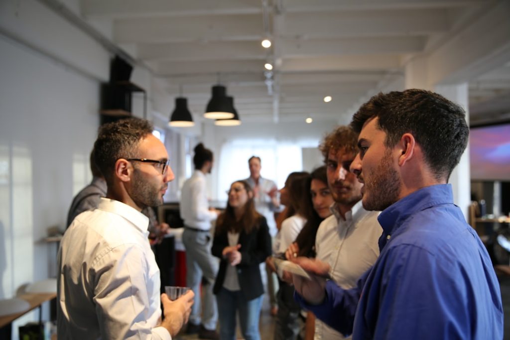 Two professionals in a focused one-on-one conversation at a business networking event, with a small group talking in the background